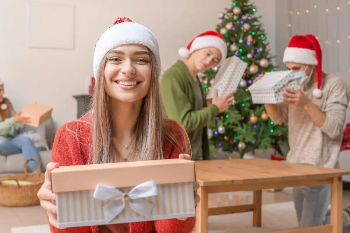 A smiling young woman in a Santa hat holding a wrapped Christmas gift with friends and a decorated tree in the background.