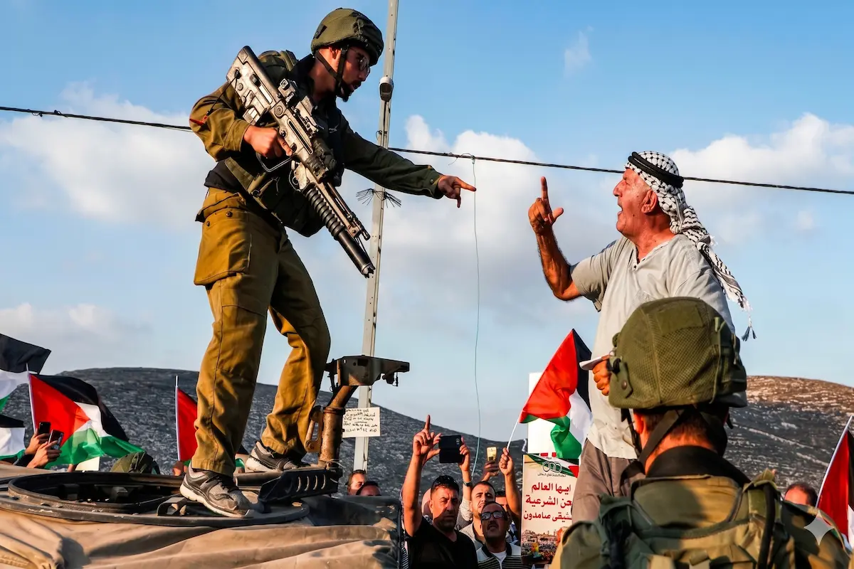 An Israeli soldier in uniform standing on an armored vehicle points at an elderly Palestinian man in a keffiyeh shouting, with flags and a crowd, during a protest, illustrating the ongoing Israel–Palestine conflict.